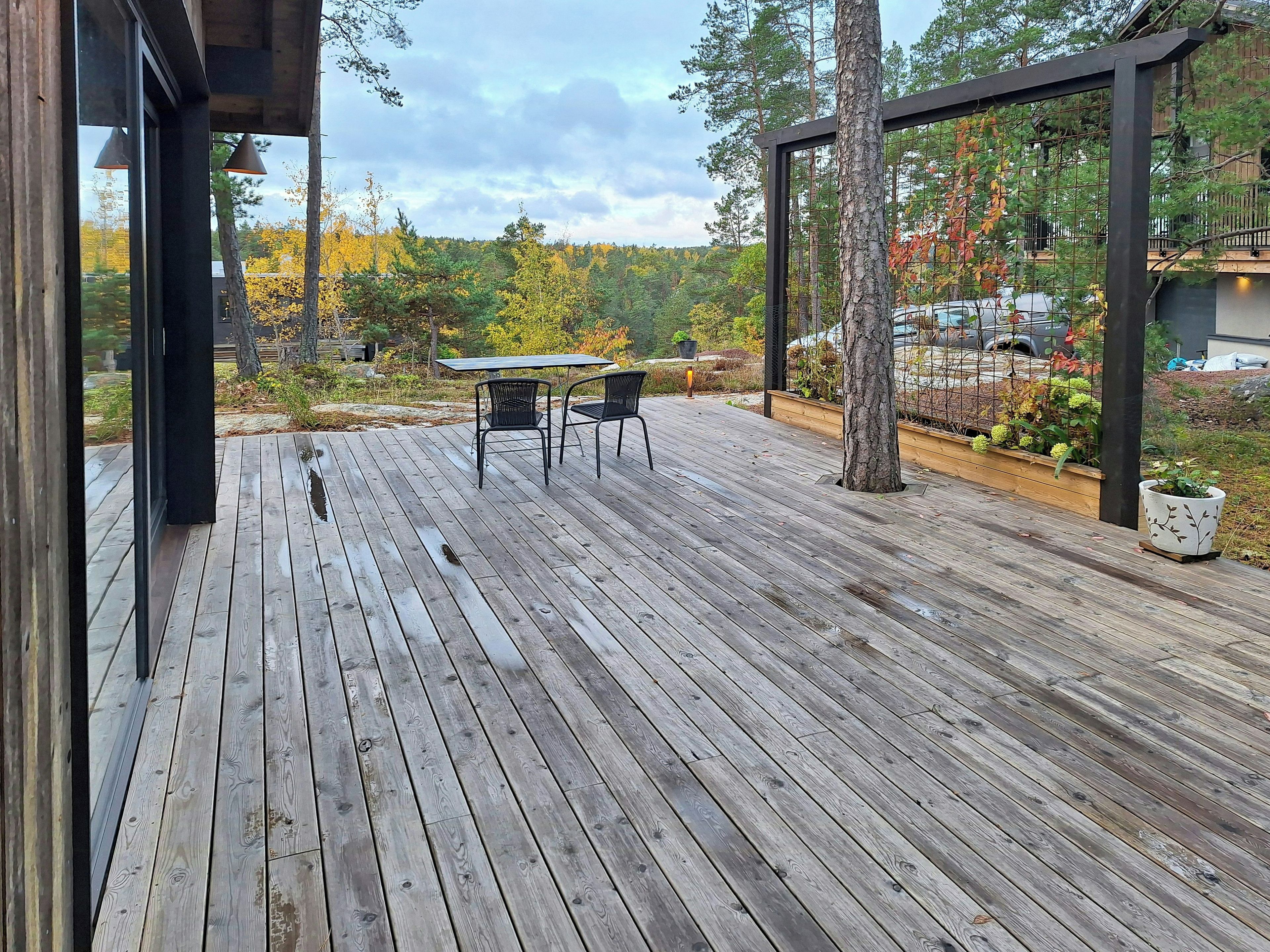 Eine Holzterrasse mit Tisch und Stühlen, umgeben von Bäumen und Blick auf einen Wald.