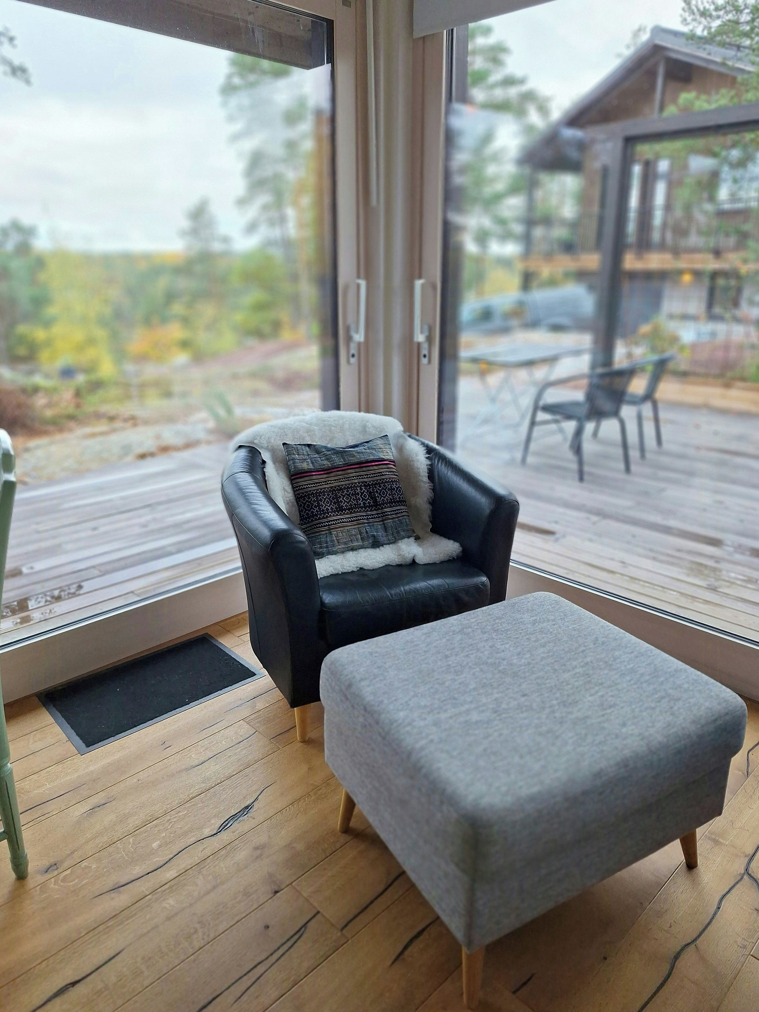 Quarto moderno com poltrona de couro preta, um banquinho cinza e uma janela grande com vista para um deck e área externa.