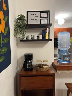 Una zona de cocina con cafetera, tazas, dispensador de agua y un cartel enmarcado con los horarios de registro de entrada y salida.