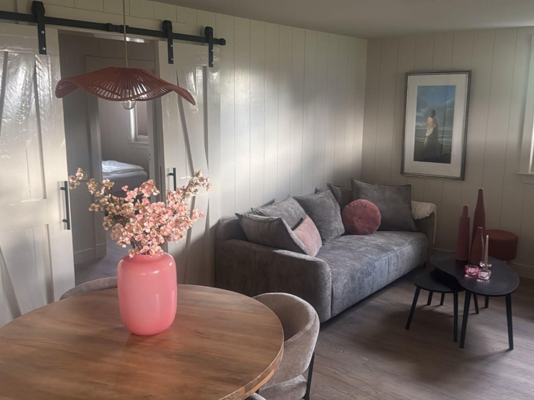 Un salón con un sofá, una mesa con un florero, un cuadro en la pared y una puerta con sistema deslizante.