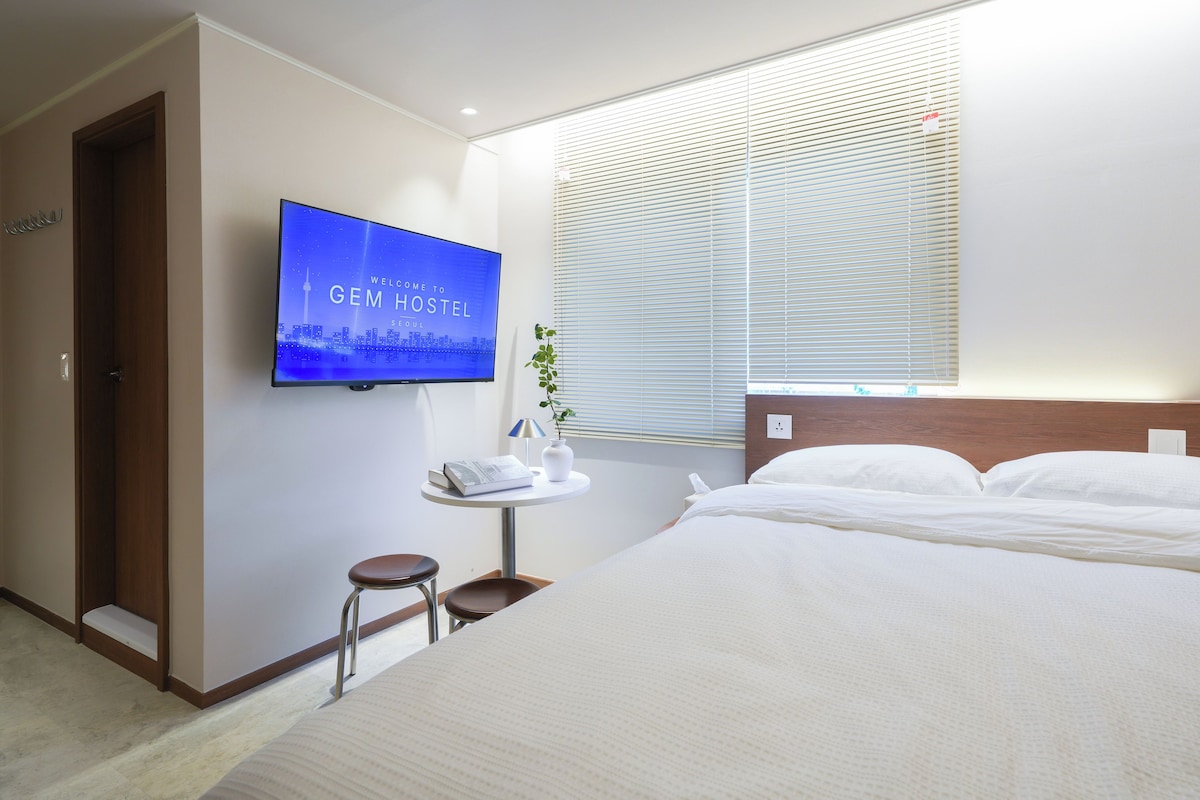 A hotel room with a bed, a bedside table with a lamp, a TV displaying a cityscape, and a window with blinds.