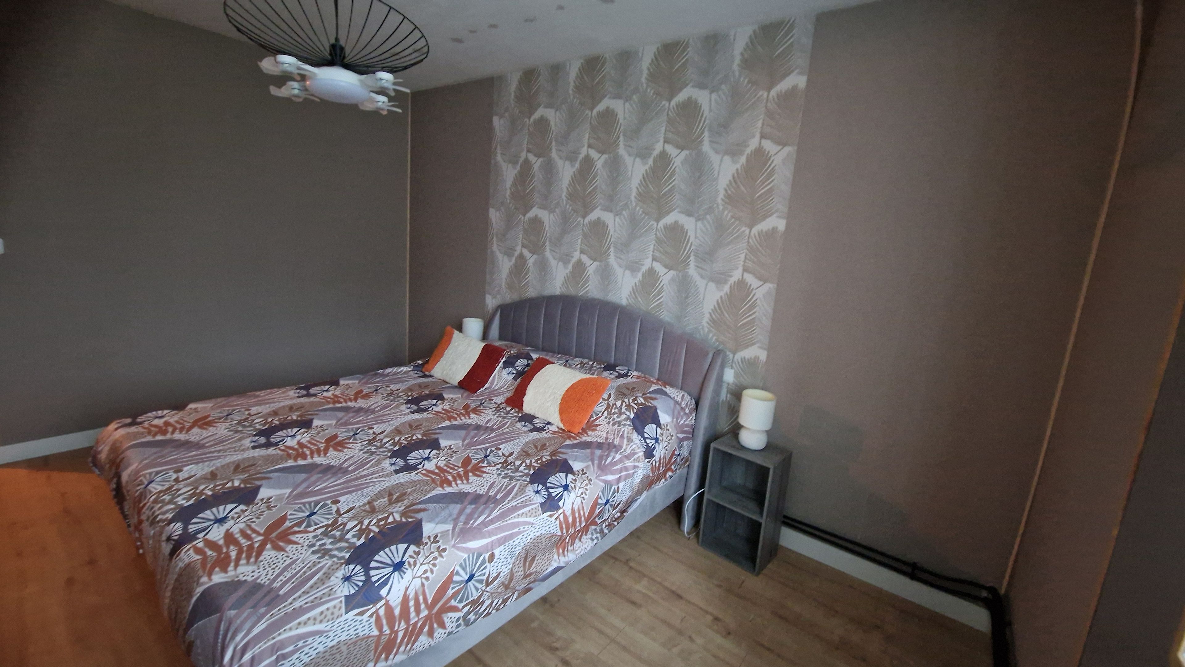 A bedroom with a bed, a bedside table, a wall-mounted fan, and a patterned wallpaper.