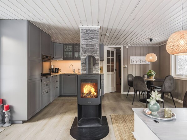 A modern kitchen with a central wood-burning stove, grey cabinets, and a dining area with a table and chairs.