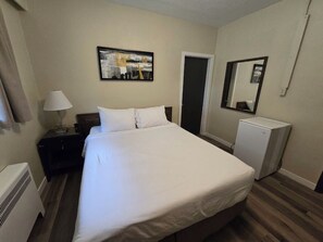 A hotel room with a bed, a nightstand, a lamp, a refrigerator, a painting on the wall, and a door. - New Charles Hotel (Boston Bar)
