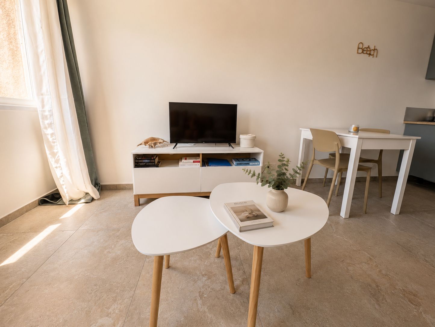 Een moderne woonkamer met een flatscreen televisie, een witte console tafel met boeken, een klein rond tafeltje met een plant en een eethoek met een stoel.