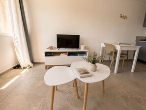 A modern living room with a flat-screen TV, a white console table with books, a small round table with a plant, and a dining area with a chair. - New Listing (Saint-Florent)