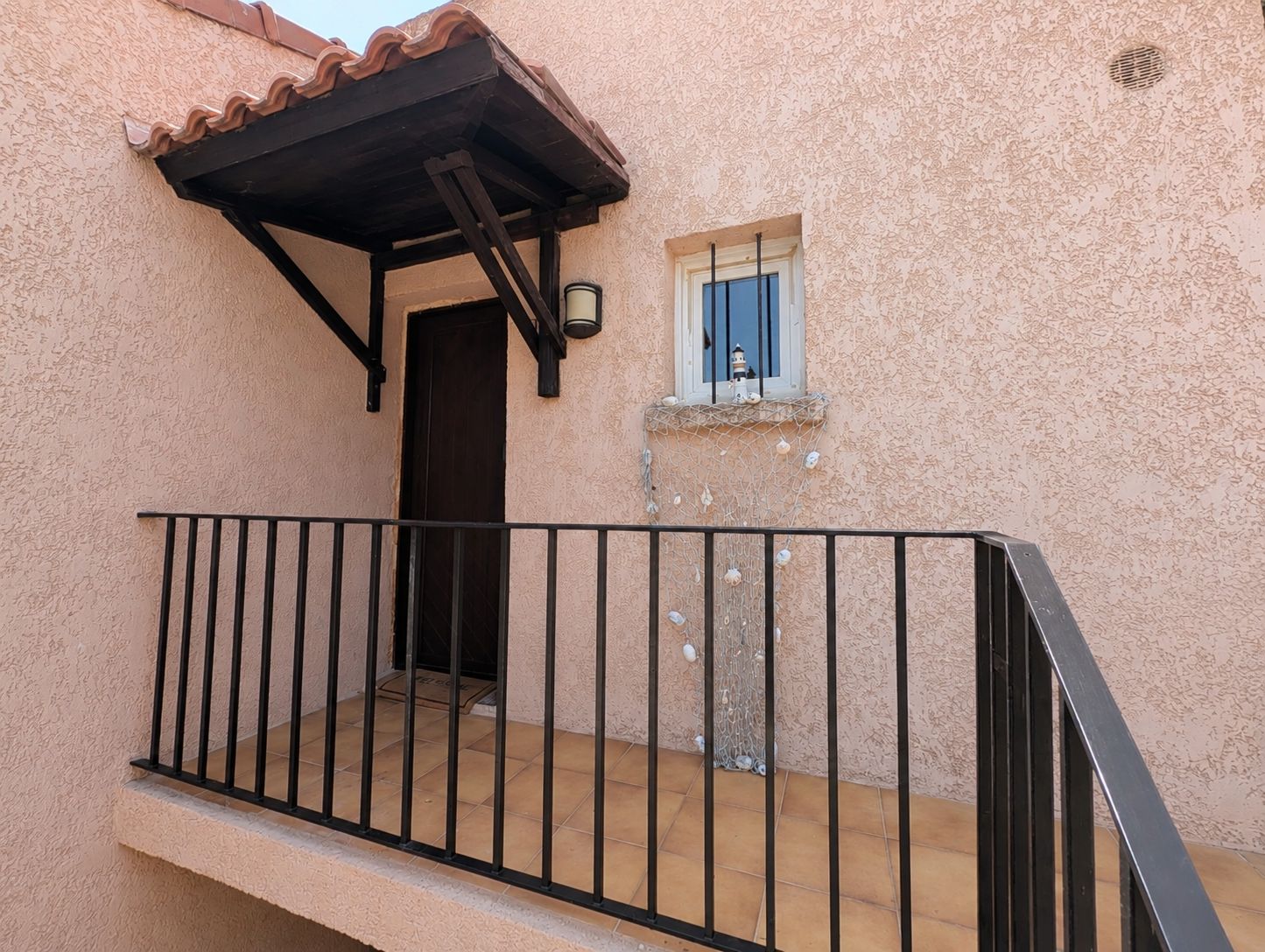 Un balcon doté d’une balustrade en métal noir, une petite fenêtre avec un volet bleu et une porte donnant sur un espace intérieur.