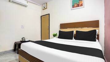 Een hotelkamer met een bed, nachtkastje met waterkoker en bekers, een wandairconditioner, een deur en een lijst met een afbeelding aan de muur.