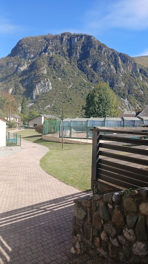 Ein gepflasterter Weg führt zu einer Grünfläche, im Hintergrund ist eine Berglandschaft zu sehen.