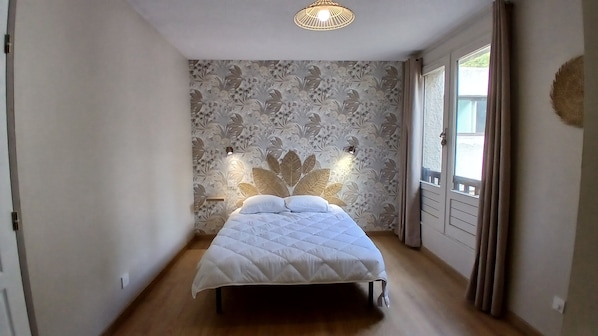 Ein Schlafzimmer mit einem Bett, blumigem Wandpapier, einer Deckenleuchte und einem Fenster mit Vorhängen.