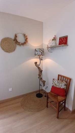 Ein Zimmer mit Holzboden, einem runden Gewebeteppich, einem hölzernen Stuhl mit rotem Kissen, einer Stehlampe und Wanddekorationen, darunter ein Kranz und ein Regal mit einem roten Gegenstand.