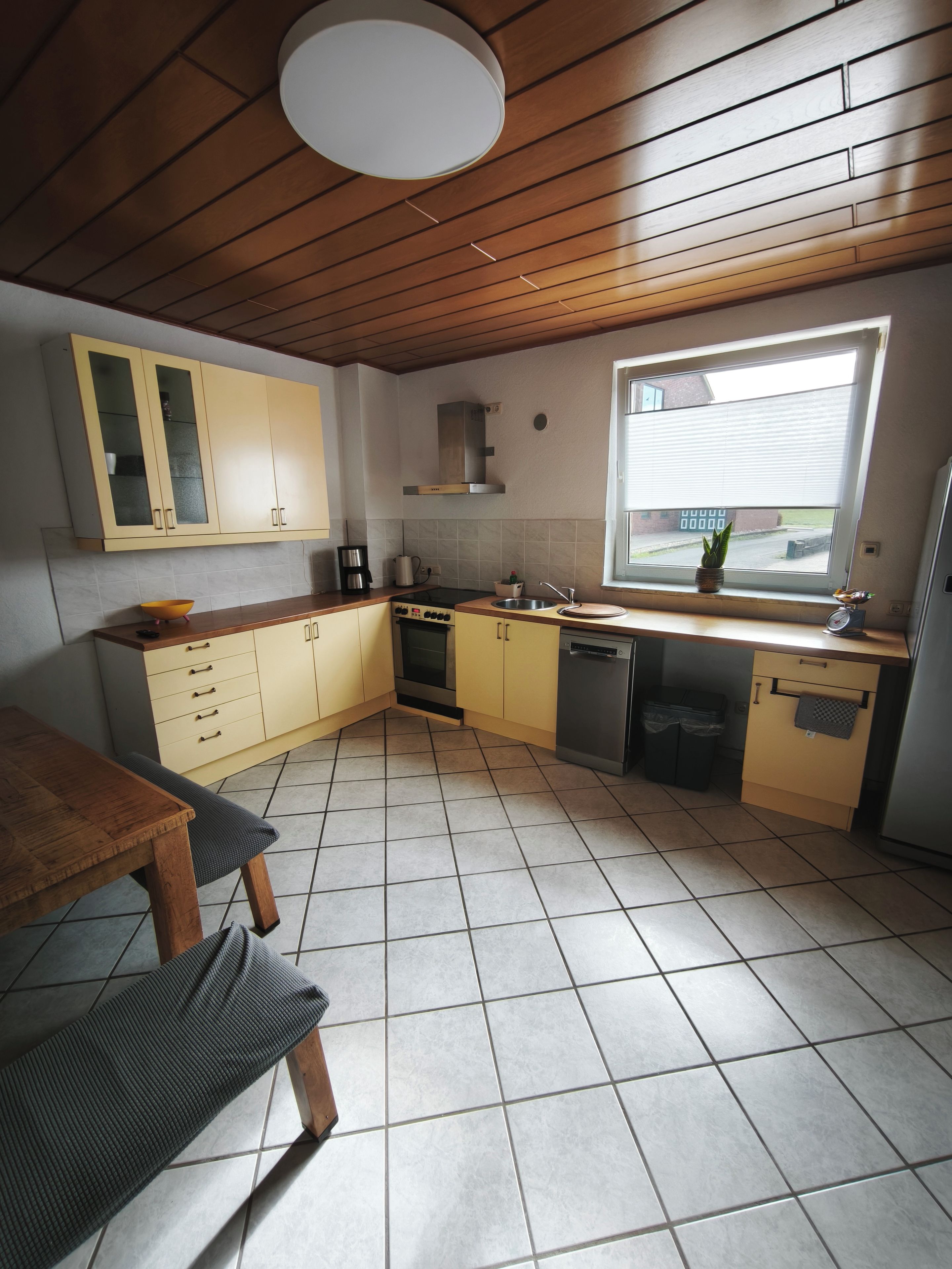 Een keuken met een houten plafond, gele kastjes, een raam en een betegelde vloer.