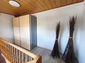 A room with a wooden ceiling, a wooden staircase railing, and two bundles of twigs against the wall.