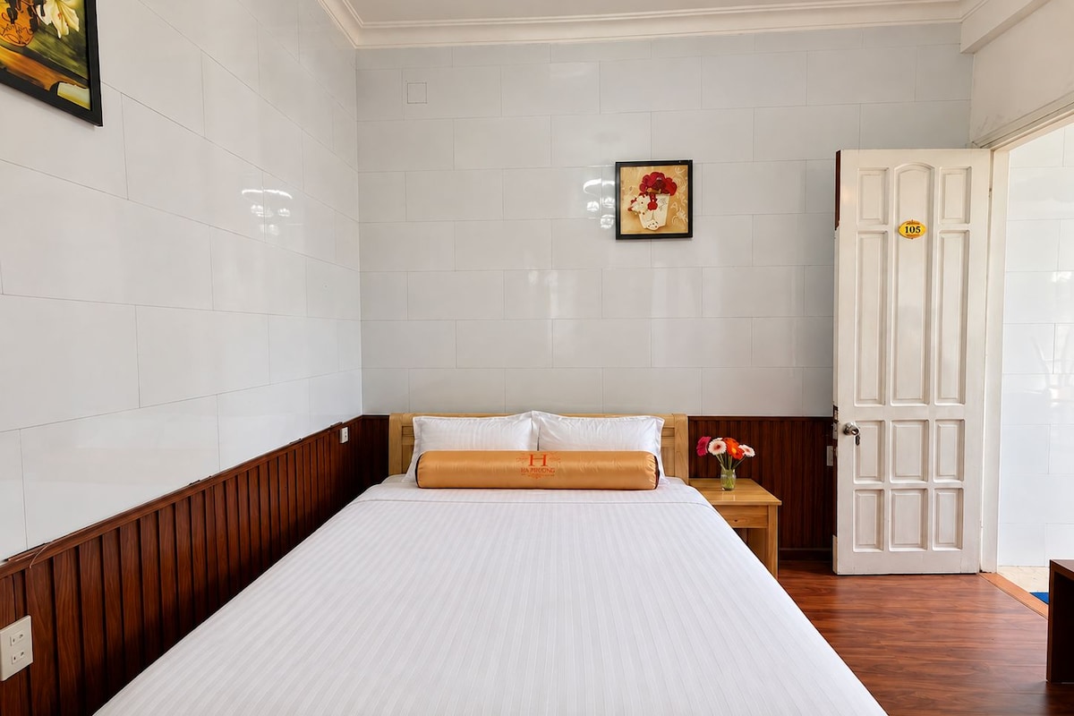 Quarto de hotel com uma cama grande, cabeceira de madeira, parede branca, porta com número e uma pequena mesa com um vaso.