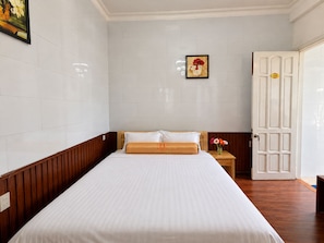 Quarto de hotel com uma cama grande, cabeceira de madeira, parede branca, porta com número e uma pequena mesa com um vaso.