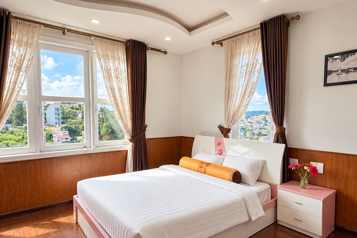 Un dormitorio con cama, mesita de noche, ventana con cortinas y vista a edificios.
