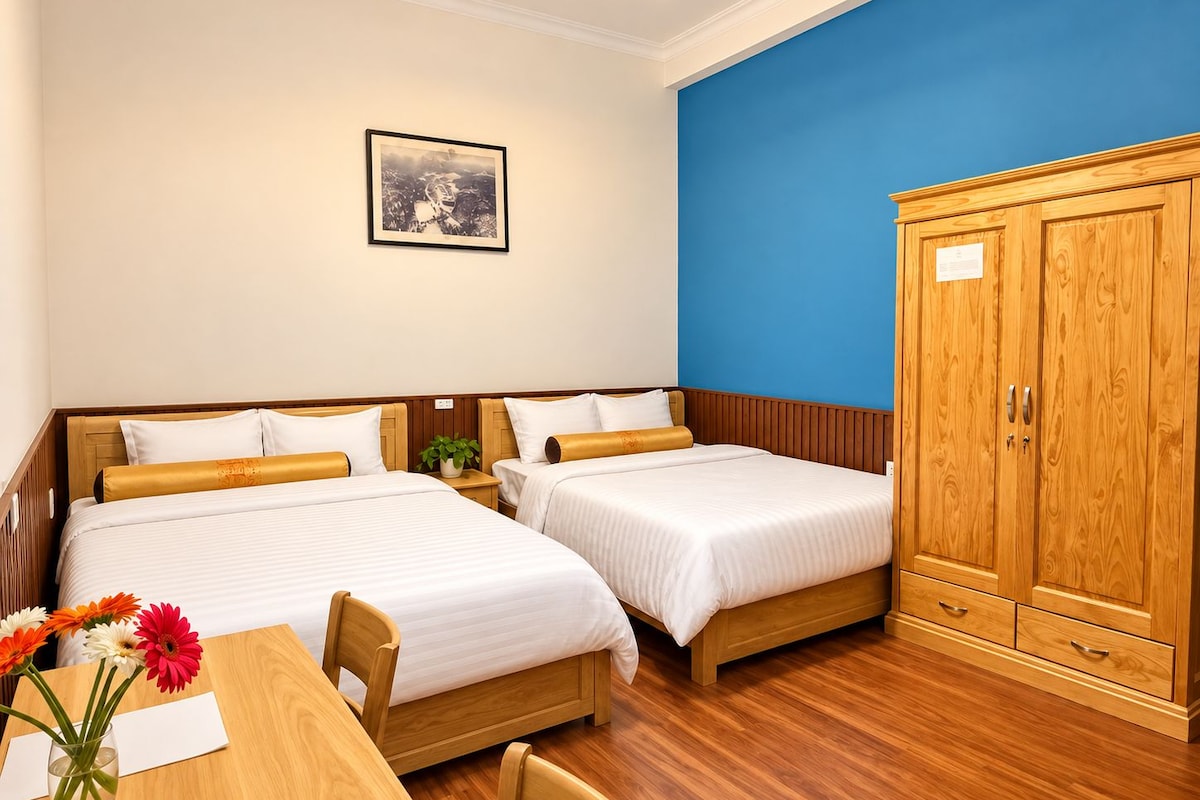 Habitación de hotel con dos camas, un armario de madera, un cuadro en la pared y una mesita con un jarrón de flores.