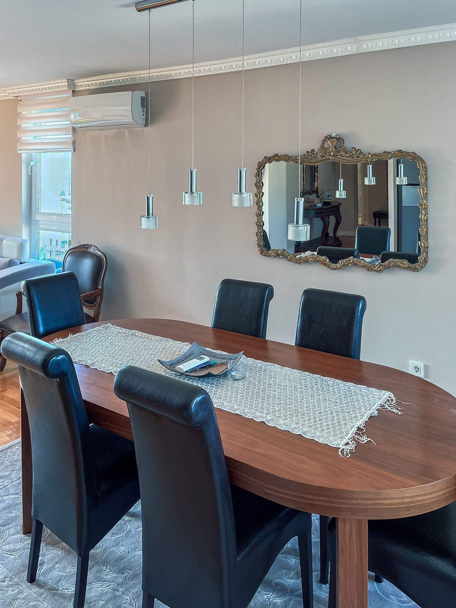 A dining area with a wooden table, black chairs, a lace table runner, pendant lights, and a large ornate mirror on the wall.