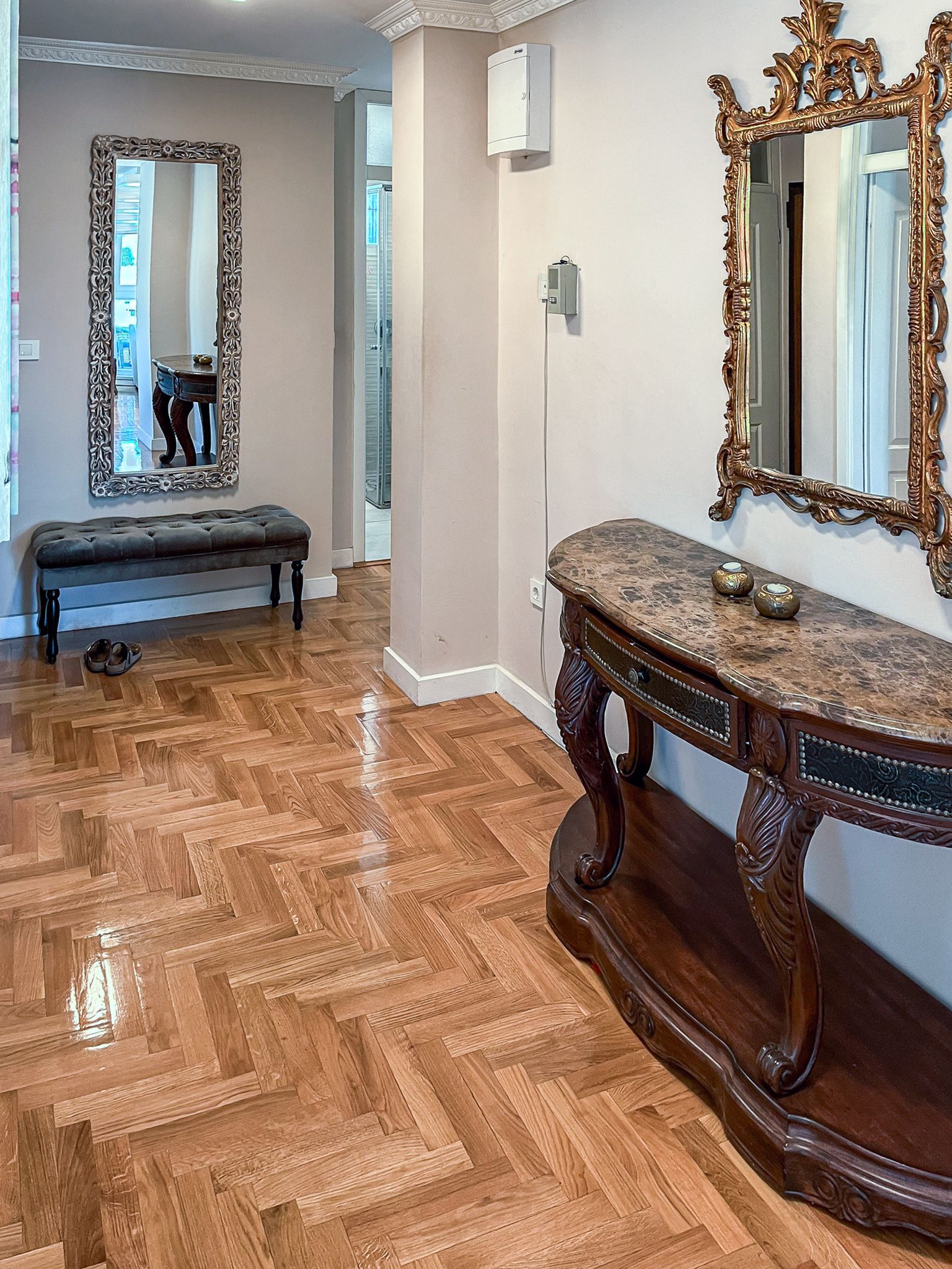 Una habitación con suelo de madera en espiga, un banco, una consola de mármol y dos espejos decorados.
