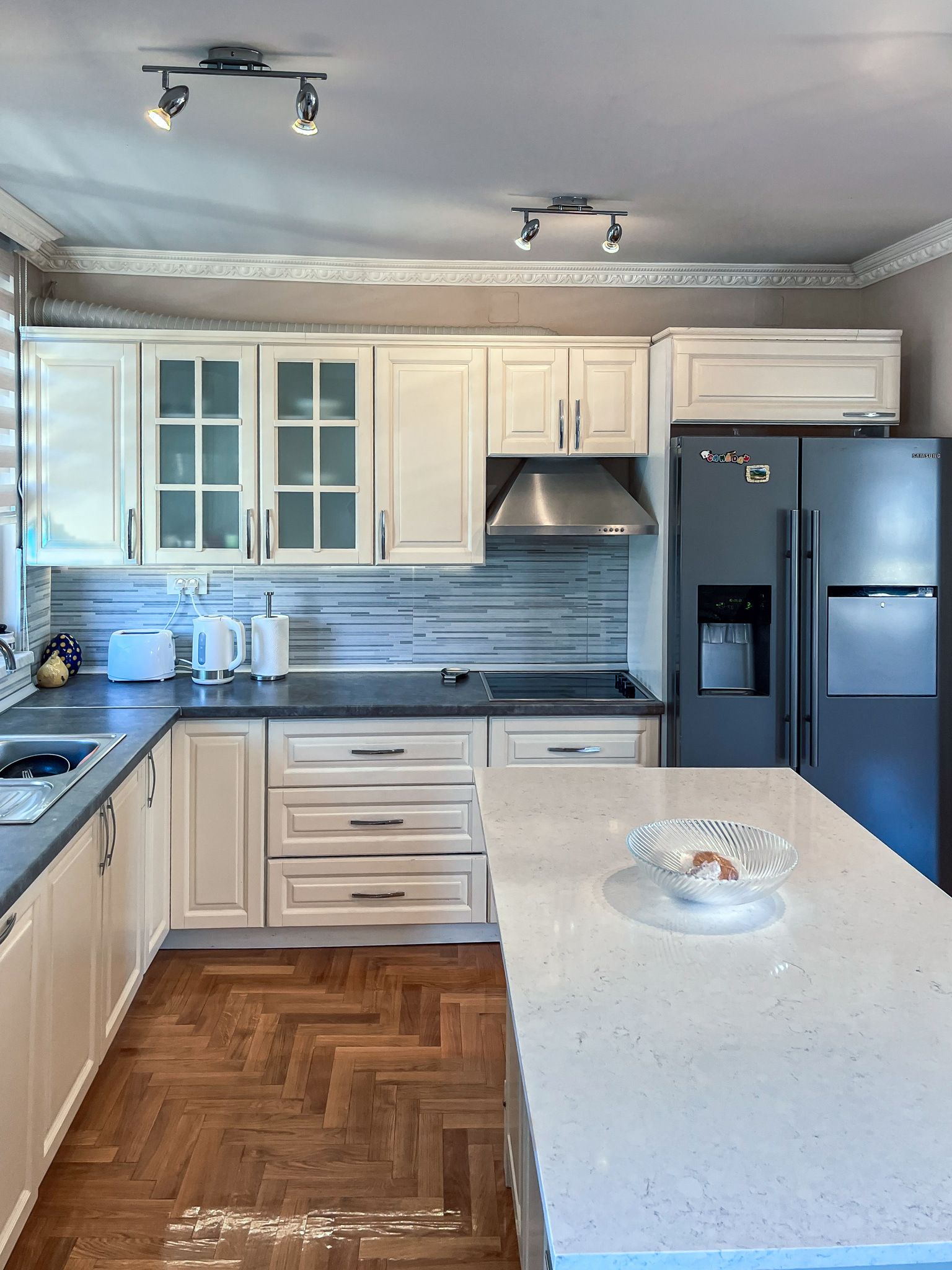 A modern kitchen with white cabinetry, a stainless steel range hood, a double-door refrigerator, and a herringbone-patterned wood floor.