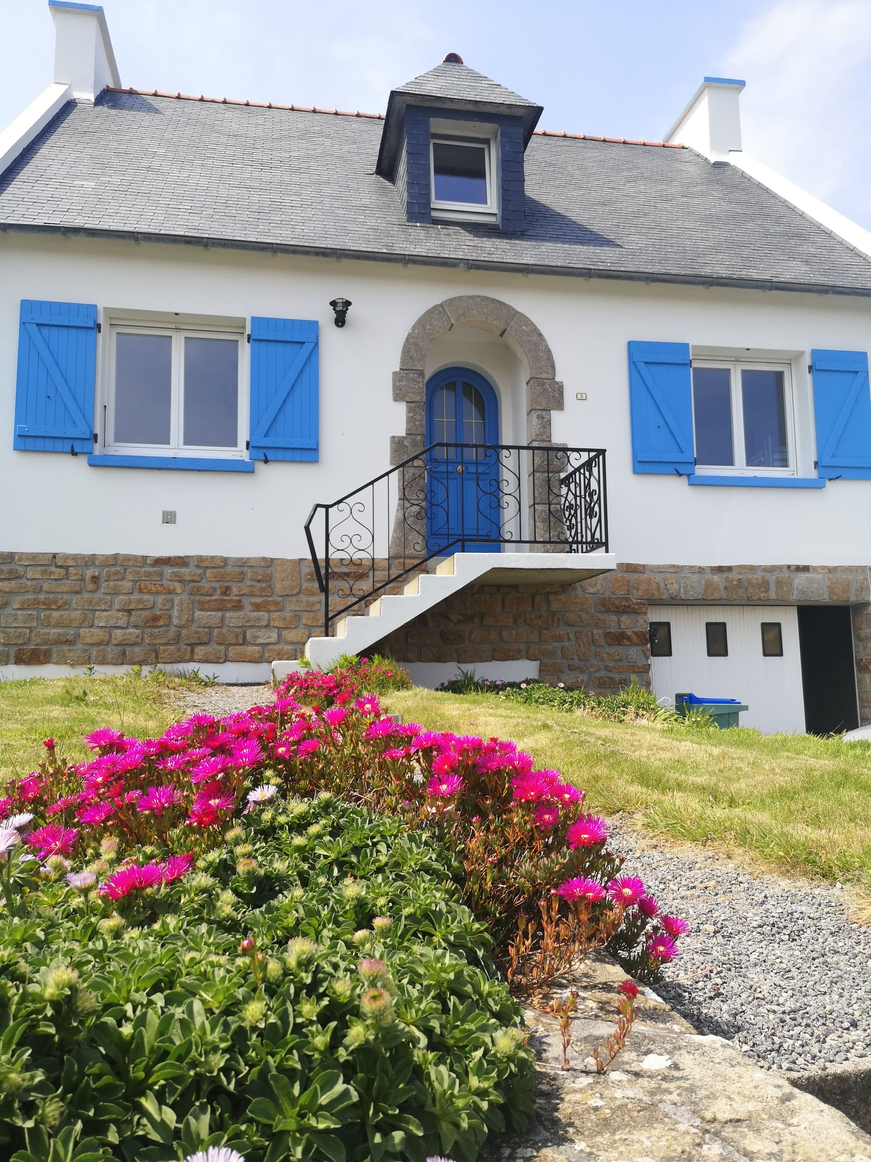 Une maison à deux étages, avec des fondations en pierre, une porte bleue et un petit balcon doté d’une balustrade.