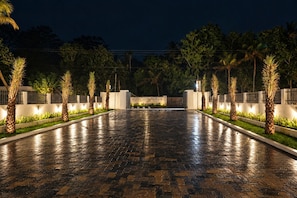 A paved pathway flanked by palm trees and illuminated by ground lights, leading to a gated entrance.