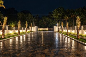 Un camino pavimentado bordeado de palmeras e iluminado con luces empotradas, que conduce a una entrada con portón.