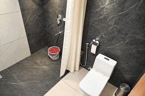 A bathroom with a toilet, shower, and a bucket of red bath items. - Hotel Legacy Heights (Padinjare Vemballur)