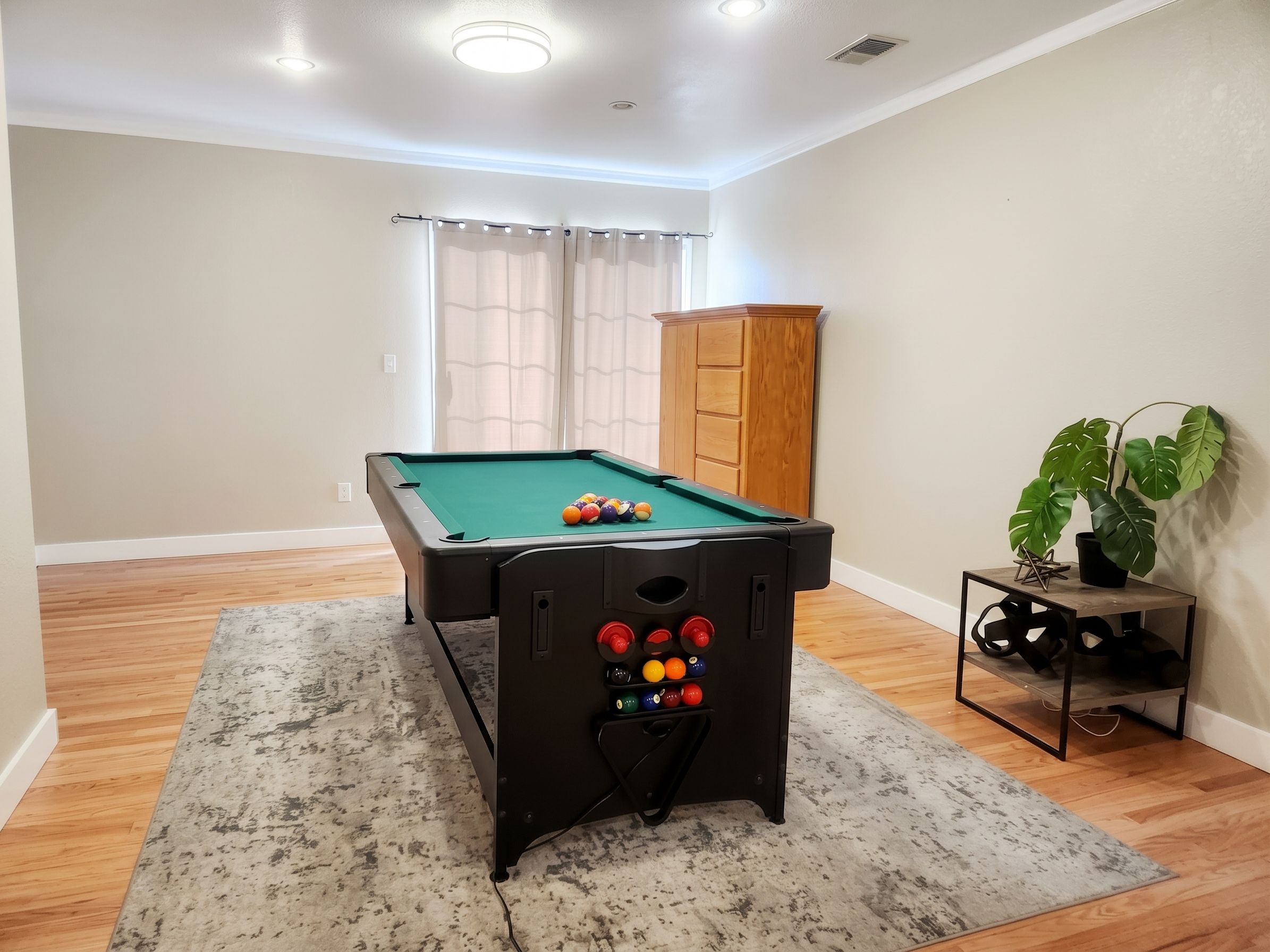 Une table de billard avec des billes, un meuble, une plante et un porte-bouteilles dans une pièce au plancher en bois et dotée d’une fenêtre avec des rideaux.