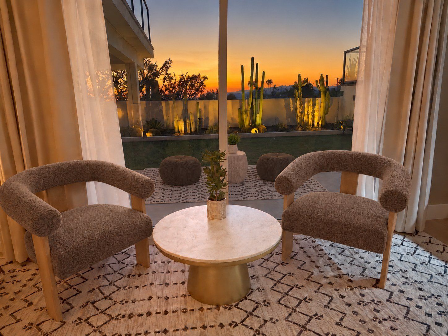Une pièce avec deux fauteuils à motifs, une table ronde et une vue sur une piscine bordée de cactus, au coucher du soleil.