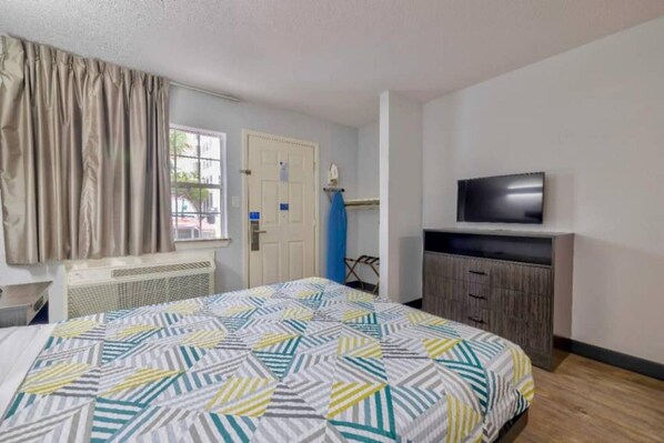 A hotel room with a bed, a television on a stand, a window with curtains, and a door. - 1 Queen Bed | ADA/RI Shower/Smoking/Kitchen (Dallas)
