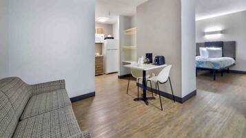 A modern hotel room with a sofa, a dining table with chairs, a kitchenette, and a bed with a headboard.