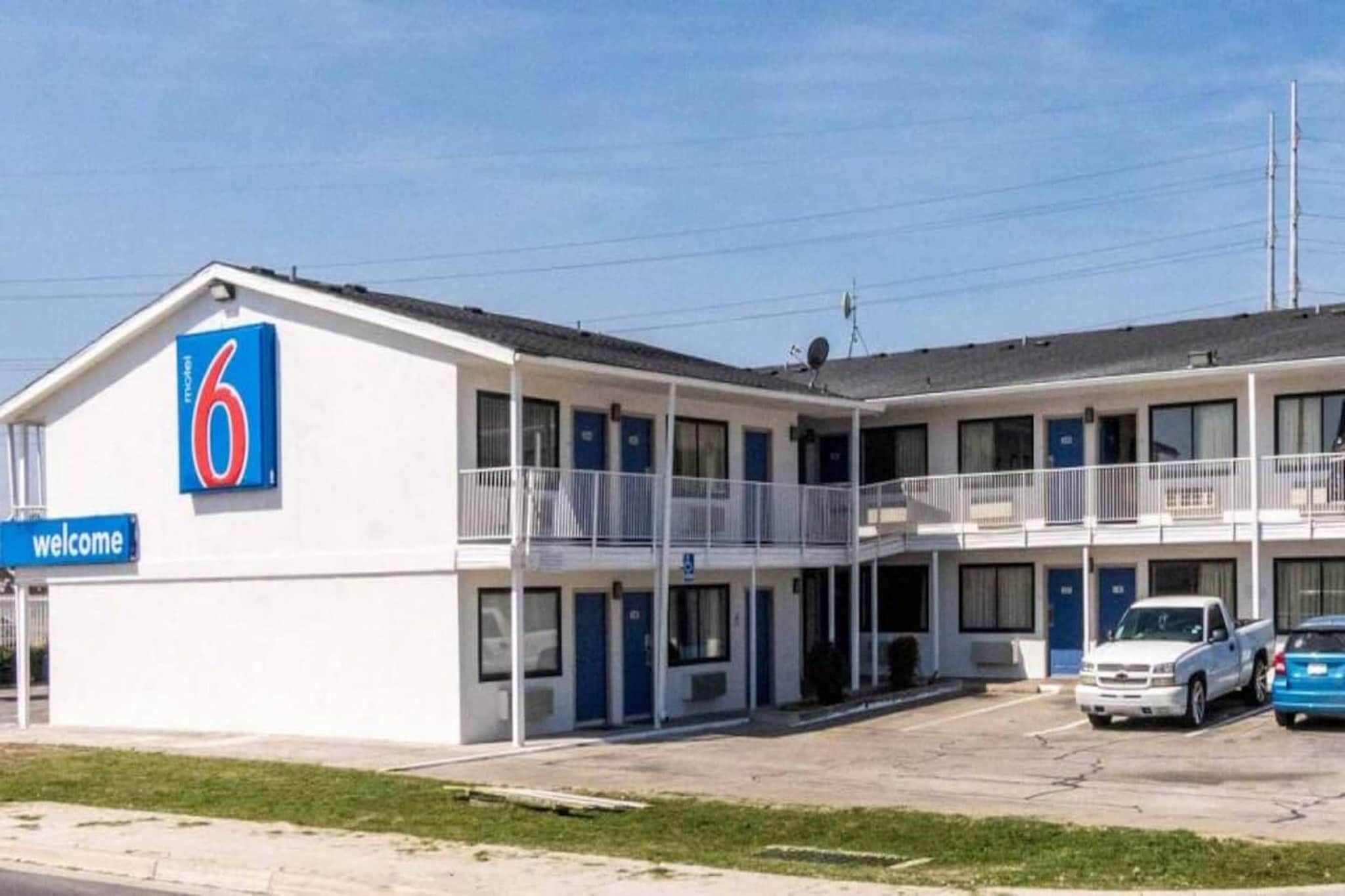 A motel with a '6' sign, a 'welcome' sign, and a parking lot with two cars.