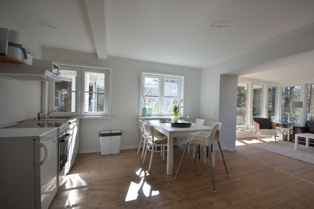 A modern kitchen with white cabinetry, a dining area with a white table and chairs, and large windows allowing natural light.