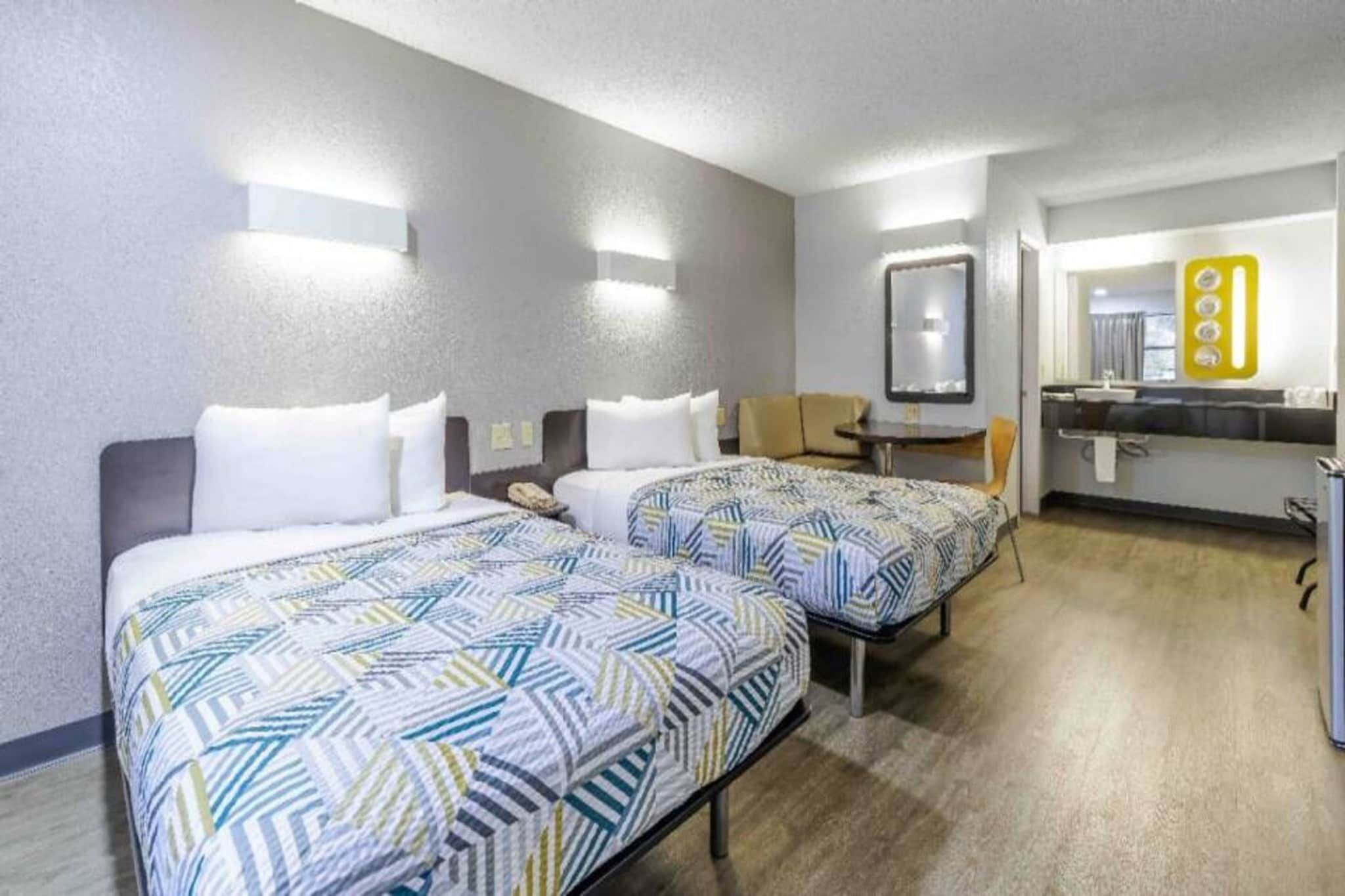 Habitación de hotel con dos camas, una mesita, una silla, un espejo y una kitchenette.