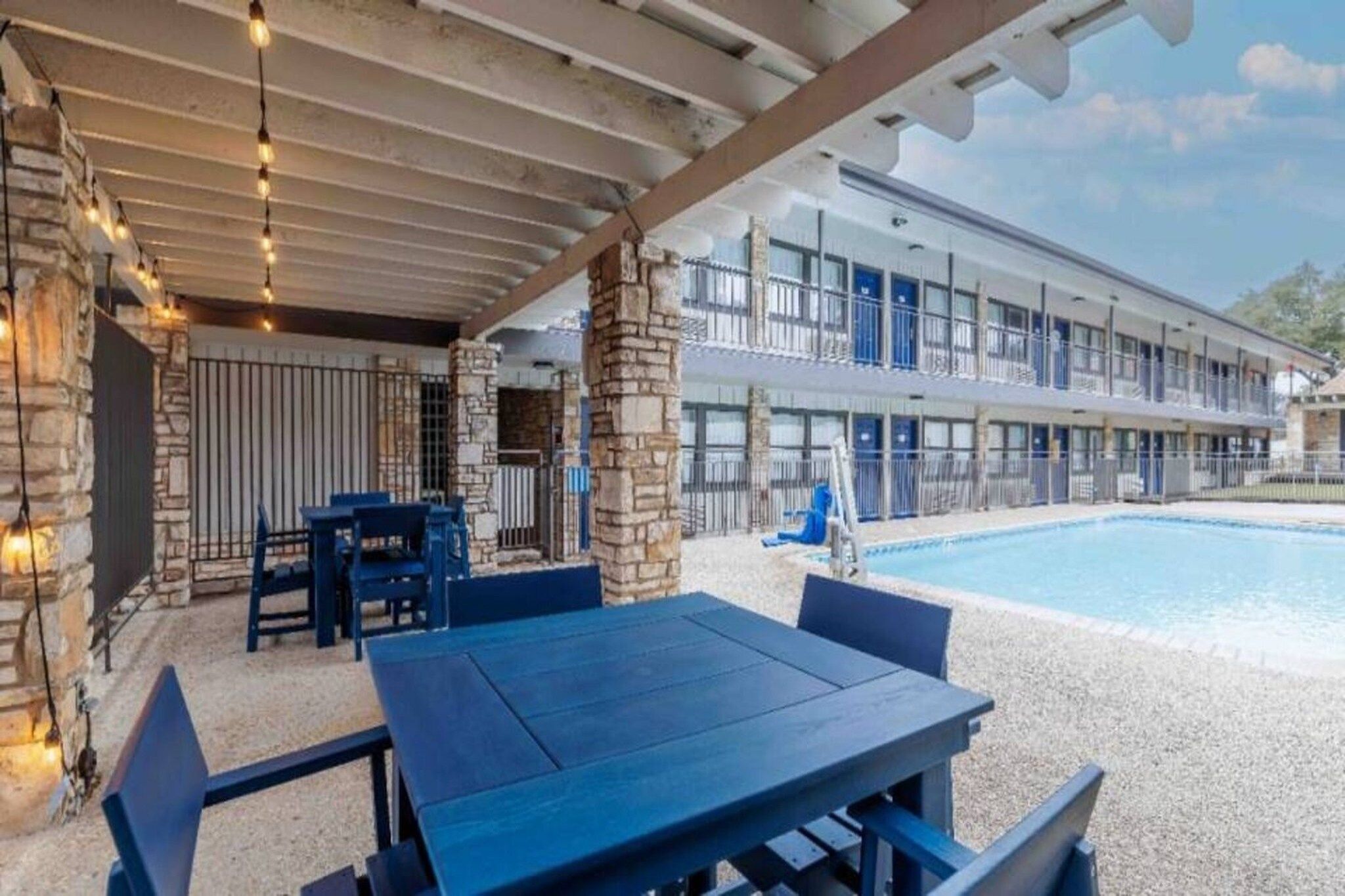 Un espace piscine extérieur doté d’un coin salon couvert, de piliers en pierre et d’un bâtiment avec de nombreux balcons en arrière-plan.