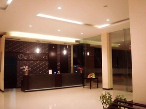 Mostrador de recepción con una encimera oscura y un cartel que dice 'De Resort'. A la derecha, una pequeña mesa con objetos y una planta en maceta.