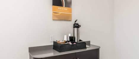 Un petit coin cuisine équipé d’une machine à café, d’un plateau noir contenant des ustensiles et d’une affiche accrochée au mur.
