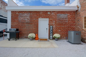 A brick building with a white door, a barbecue grill, two potted plants, a welcome sign, and an air conditioning unit. - Galena Route 66 Retreat w/ Historic Brick Style (Galena)