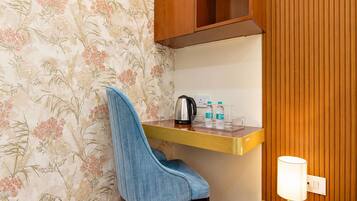 A room with a floral wallpaper, a blue upholstered chair, a wooden cabinet, a small table with a kettle, bottles, and a lamp.