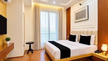 A modern hotel room with a large bed, a bedside table with a lamp, a mounted TV, and a window with curtains.