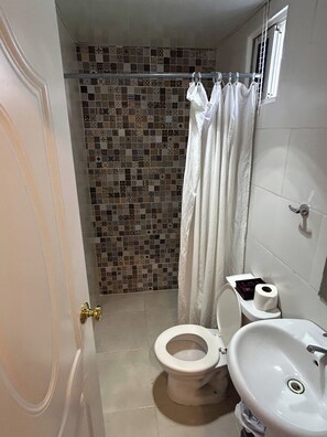 A bathroom with a tiled shower wall, a toilet, a sink, and a window with curtains. - Blue Night Aparta Hotel Your haven of peace (Punta Cana)
