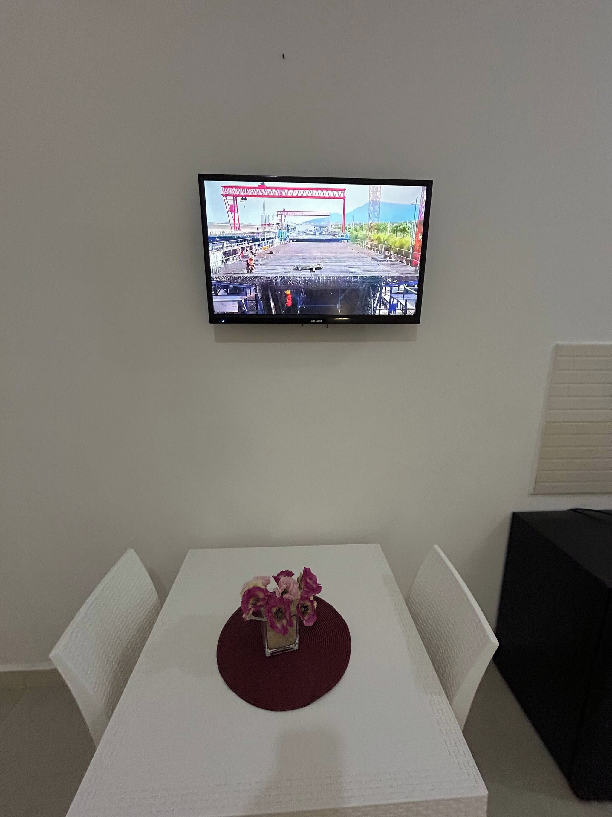 Een aan de muur bevestigde televisie toont een bouwterrein. Daaronder staat een witte tafel met een rode placemat en een bloevas.