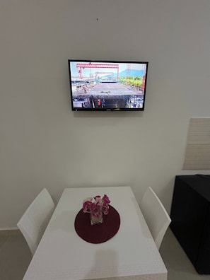 Un televisor montado en la pared muestra una obra de construcción. Abajo, una mesa blanca con un mantel individual rojo y un jarrón de flores es visible.