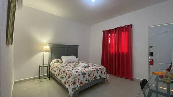 Un dormitorio con una cama, una mesita de noche con lámpara, una cortina roja y una puerta.