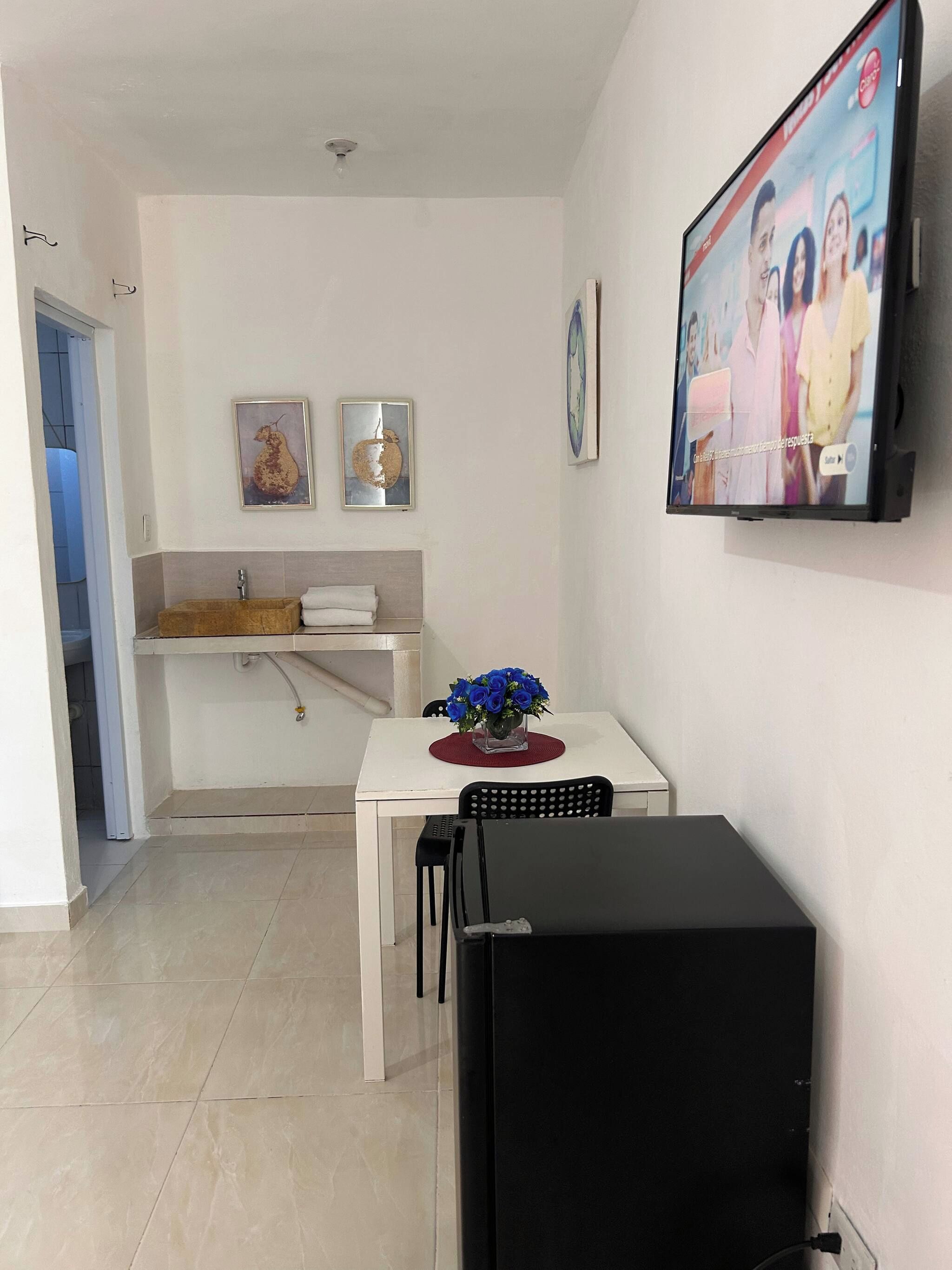 Een gezellig compact leefruimte met een kitchenette, een eethoek met tafel en stoelen, een zwart kastje en een aan de muur bevestigde televisie.