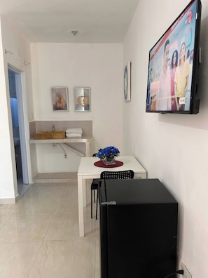 Un espai vital compacte amb una petita cuina, una zona de dinar amb taula i cadires, un armari negre i un televisor fixat a la paret.