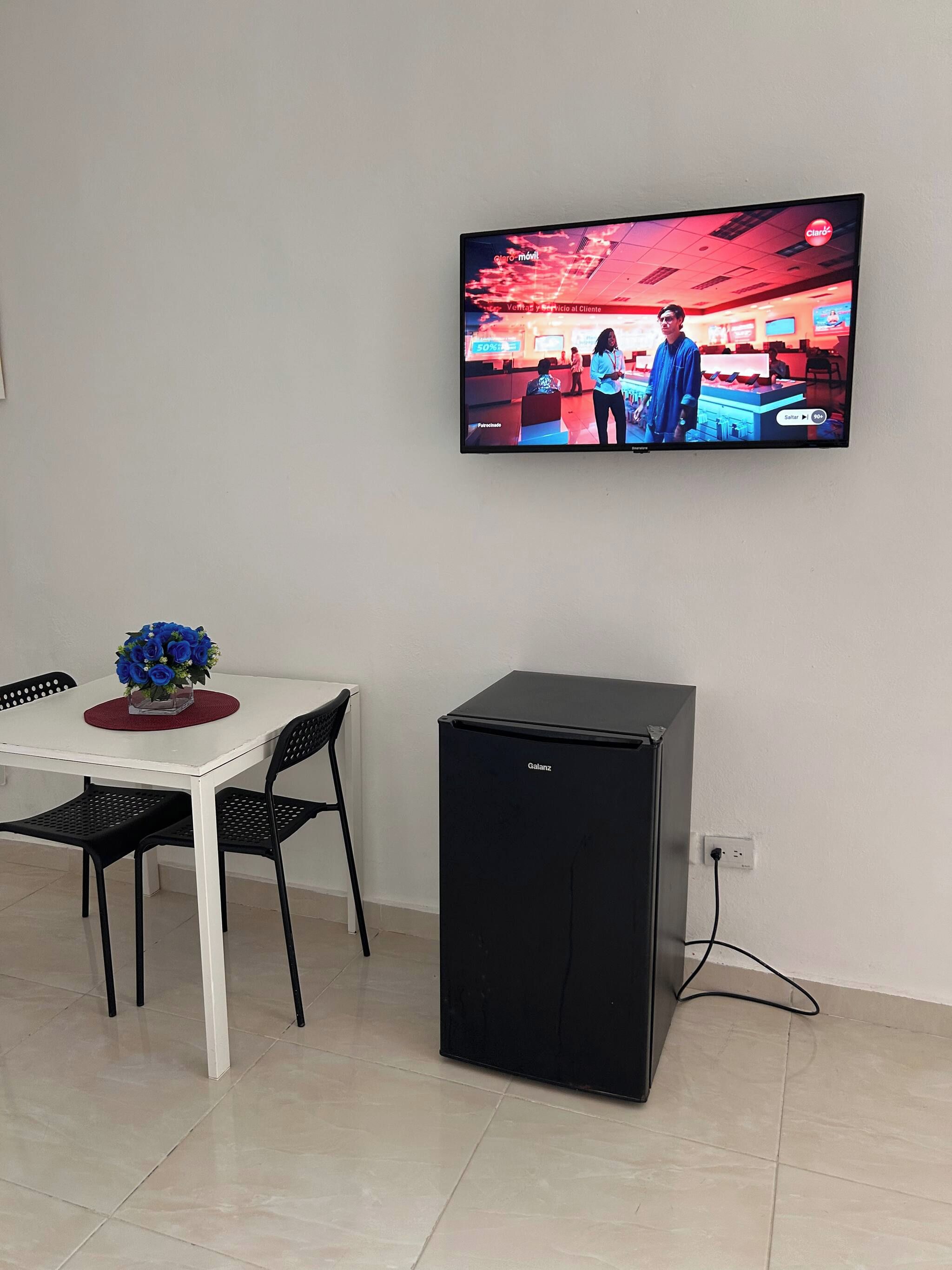 Een kamer met een aan de muur bevestigde televisie, een kleine, zwarte koelkast, een witte tafel met een bloevas en twee zwarte stoelen.