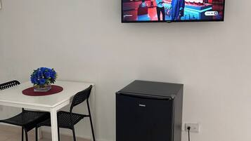 Een kamer met een aan de muur bevestigde televisie, een kleine, zwarte koelkast, een witte tafel met een bloevas en twee zwarte stoelen.