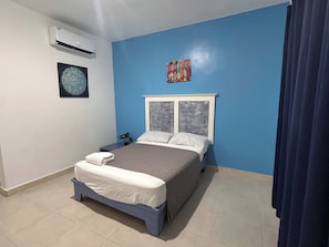 Un dormitorio con una cama, una mesita de noche, aire acondicionado de pared y una ventana con cortinas.